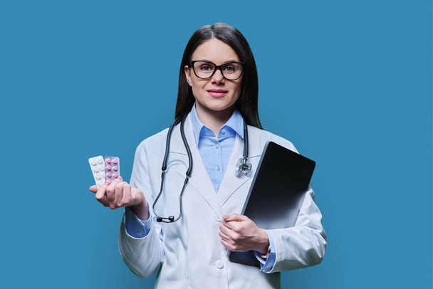 Young woman doctor recommending different pills in blister pack, female smiling holding laptop looking at camera on blue background. Medicine, healthcare, treatment, pharmacy, pharmacology concept - Fotó, kép