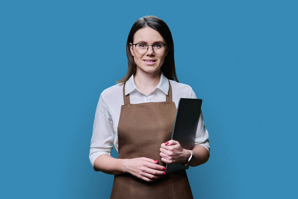 Portrait of young worker woman in apron holding laptop on blue color studio background. Confident smiling female looking at camera, service worker, small business owner, salesperson, startup concept - Photo, Image