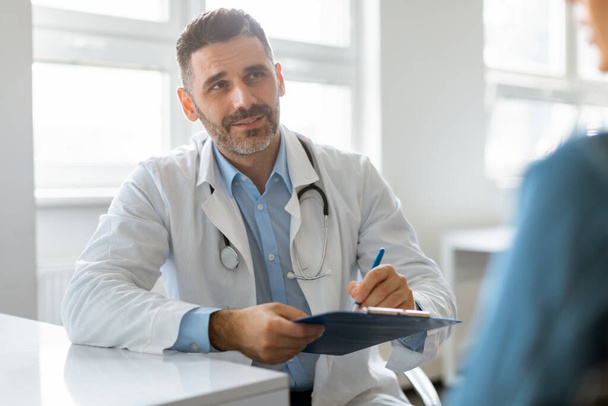 Portrait of cheerful middle aged male doctor listening to female patient, discussing diagnosis and treatment, writing in clipboard. Specialist giving consultation to lady - Fotoğraf, Görsel
