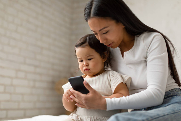 Azjatycka mamusia i mała córeczka za pomocą smartfona oglądanie kreskówek online i Websurfing siedzi na łóżku w nowoczesnej sypialni w domu. Koncepcja dzieci i gadżetów. Skupienie selektywne - Zdjęcie, obraz