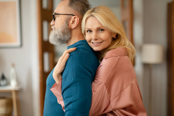 Szczęśliwa dojrzała żona przytulająca swojego męża stojącego za jego plecami i uśmiechającego się do kamery w domu. Małżonkowie obejmujący pozowanie w nowoczesnym salonie. Szczęście w małżeństwie. Skupienie selektywne - Zdjęcie, obraz