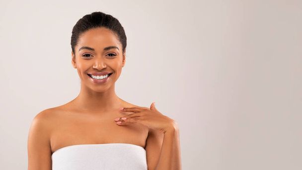 Seksownie atrakcyjna młoda czarna kobieta pozująca półnago na szarym tle pracowni, owinięta w ręcznik, dotykająca jedwabiście miękkiej skóry po prysznicu lub zabiegu SPA i uśmiechnięta, przestrzeń do kopiowania. Koncepcja pielęgnacji ciała - Zdjęcie, obraz
