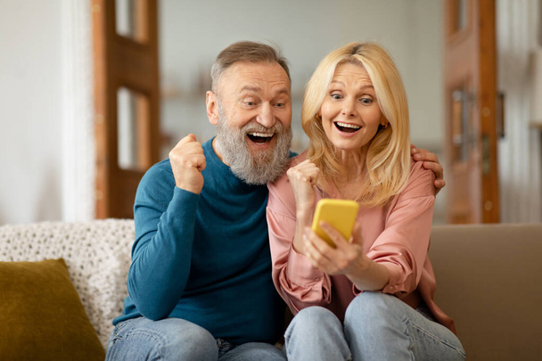 Radosna para seniorów korzystająca ze smartfona czytającego wiadomość i trzęsących się pięściami świętująca wielkie szczęście siedząc na kanapie w domu. Wow Mobile Oferta i aplikacja Reklama Koncepcja - Zdjęcie, obraz
