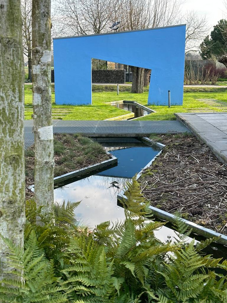 Apeltern, Hollanda, Nisan 09, 2023: Mavi duvarı, kemeri, göleti ve çimenlikteki beton yolları olan modern avangart teras. Bitkiler: eğrelti otları eritrosora, Himalaya huş ağaçları. - Fotoğraf, Görsel