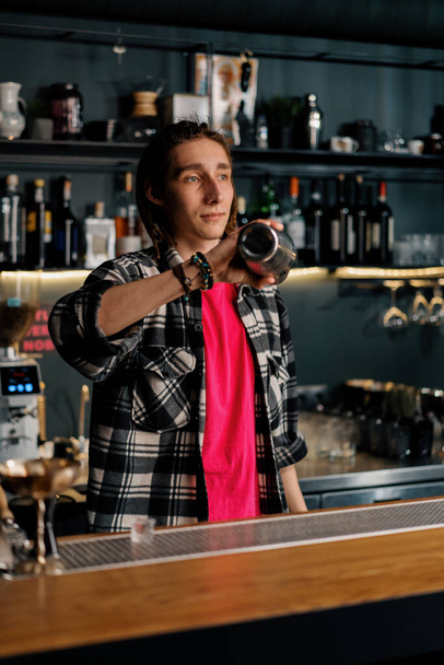 a young bartender with dreadlocks mixes the ingredients of an alcoholic cocktail by shaking shaker - Photo, Image