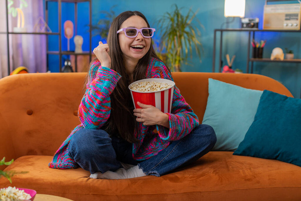 Excited woman in 3D glasses sits on couch eating popcorn snacks and watching interesting TV serial sport game, film online social media movie content at home apartment. Girl enjoying entertainment - Fotoğraf, Görsel
