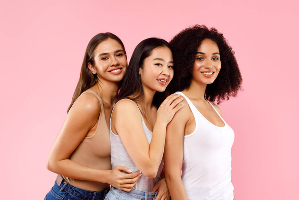 Drei wunderschöne multiethnische Millennial-Frauen, die sich umarmen und in die Kamera lächeln, stehen auf rosa Hintergrund, Studioaufnahme. Vielfältige weibliche Schönheit - Foto, Bild