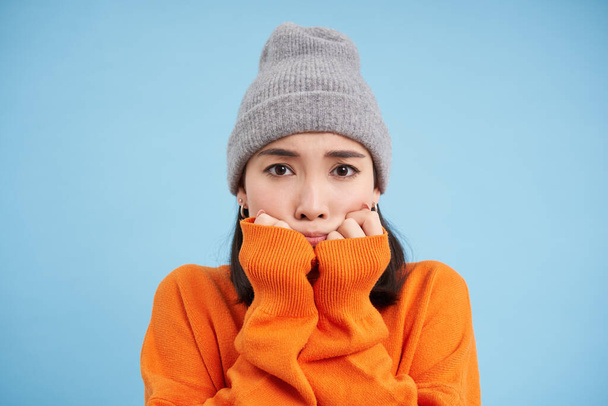 Portrait of asian girl in warm hat, looks with fear, shaking and trembling scared, standing frightened against blue background. - Foto, Imagen