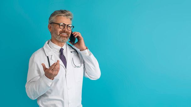 Счастливый красивый седовласый врач средних лет имеет телефонный звонок с пациентом, консультируясь дистанционно, улыбаясь и жестикулируя на фоне голубой студии, глядя на копировальное пространство, панораму - Фото, изображение