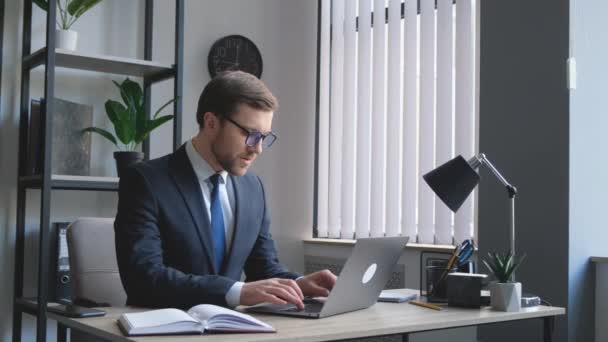 Przystojny menadżer z laptopem w biurze. Pewny siebie biznesmen siedzi przy stole biurowym i pracuje z laptopem, pisze. - Materiał filmowy, wideo