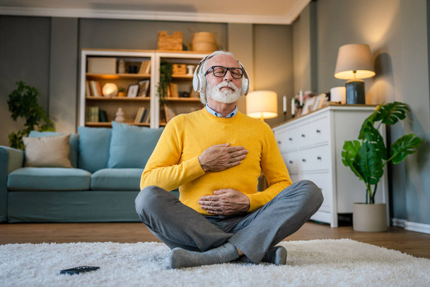 jeden mężczyzna Dojrzałe starszy kaukaski mężczyzna za pomocą słuchawek dla online prowadzony medytacja praktykuje uważność jogi manifestacja z oczami zamkniętymi w domu prawdziwe ludzie self care koncepcja kopia przestrzeń - Zdjęcie, obraz