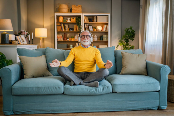 un homme mature homme caucasien aîné utilisant des écouteurs pour la méditation guidée en ligne pratiquant la pleine conscience manifestation du yoga avec les yeux fermés à la maison personnes réelles self care concept copier l'espace - Photo, image