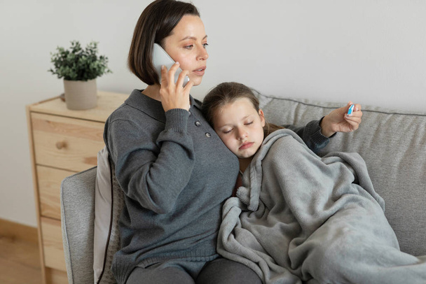 Sad worried european millennial mother calms, takes care of sick teen girl, calls by phone to doctor in living room interior. Medicine for treatment flu and cold, love, relationships at home - Fotografie, Obrázek