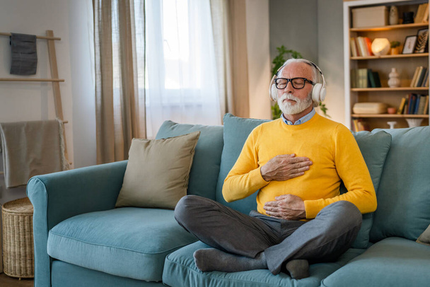 un hombre adulto mayor caucásico masculino usando auriculares para la meditación guiada en línea practicando mindfulness manifestación de yoga con los ojos cerrados en casa personas reales autocuidado concepto copia espacio - Foto, imagen
