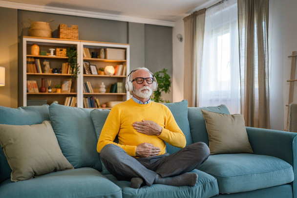 un hombre adulto mayor caucásico masculino usando auriculares para la meditación guiada en línea practicando mindfulness manifestación de yoga con los ojos cerrados en casa personas reales autocuidado concepto copia espacio - Foto, Imagen