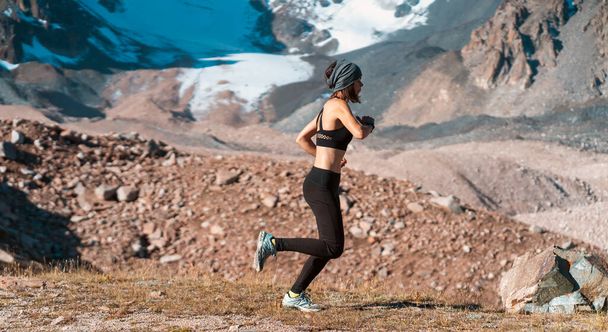 Młoda atletyczna dziewczyna uprawiająca jogging w malowniczej okolicy pośród ośnieżonych gór o zachodzie słońca. Biegacz w legginsach i top rozgrzewa się, ćwiczy i bierze udział w bieganiu szlakiem. - Zdjęcie, obraz