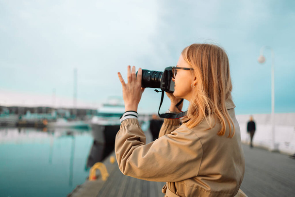 Outdoor summer smiling lifestyle portrait of pretty young woman having fun in the city in Europe in evening with camera travel photo of photographer. Making pictures on the pier near the expensive - Photo, Image