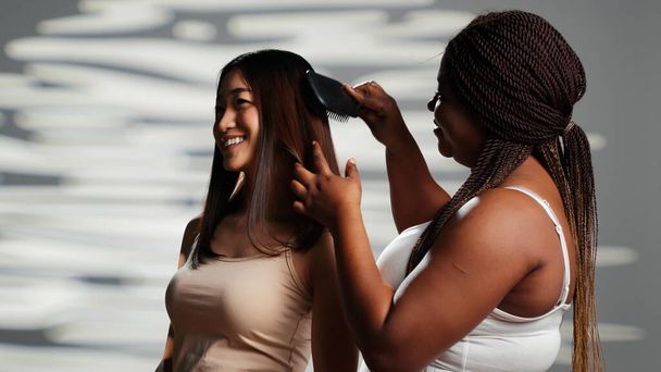 Уверенный в себе человек, расчесывающий волосы азиатской женщины, используя расческу для расчёски и выражающий концепцию принятия тела. Позитивные модели красоты, позирующие для рекламной кампании по уходу за кожей, разнообразие типов тела. - Фото, изображение