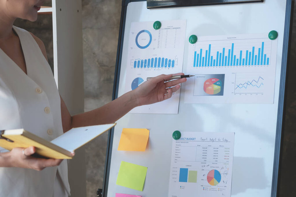 Asijská podnikatelka prezentující obchodní data s tabulí s grafy, grafy rukou drží žlutý zápisník, a obchodní dokumenty ležící na stole v kanceláři, obchodní koncept. - Fotografie, Obrázek