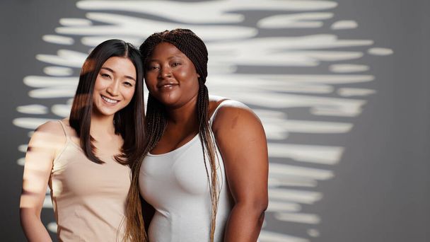 Smiling beauty models hugging and looking at camera, expressing positive emotions and bodycare. Beautiful interracial young women with different body types and skintones posing. - Photo, Image