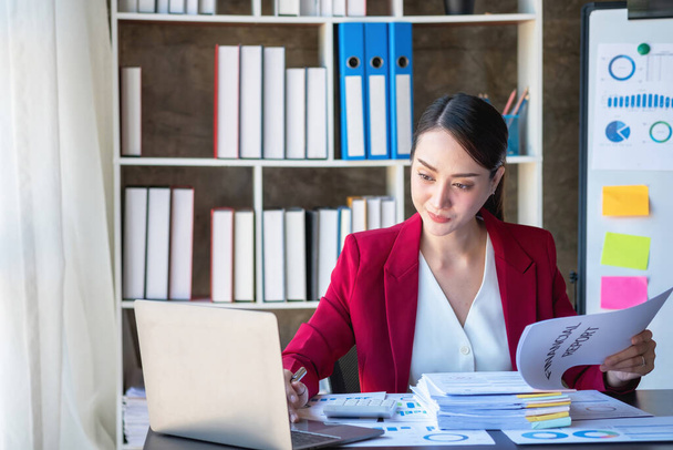 Фінансова, азійська бізнесменка в червоному костюмі, яка сидить за столом при офісі, має комп'ютер для бухгалтерського обліку на робочому місці, щоб розрахувати річний прибуток за обов'язком, бізнес ідея - Фото, зображення