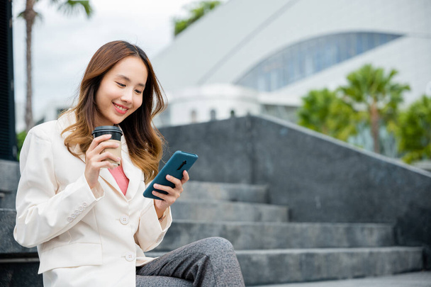 Young Asian woman wearing blue shirt while use cellphone with holding coffee cup sitting on staircase. Female business people using smart phones in the city outdoors. - Foto, imagen