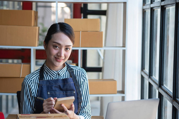 Azjatka bizneswoman cieszy się swoją działalnością internetową MŚP, sprzedając produkty, czerpiąc korzyści z otwarcia sklepu internetowego i przyjmując zamówienia na paczki klientów wewnątrz biura. Koncepcja działalności MŚP. - Zdjęcie, obraz