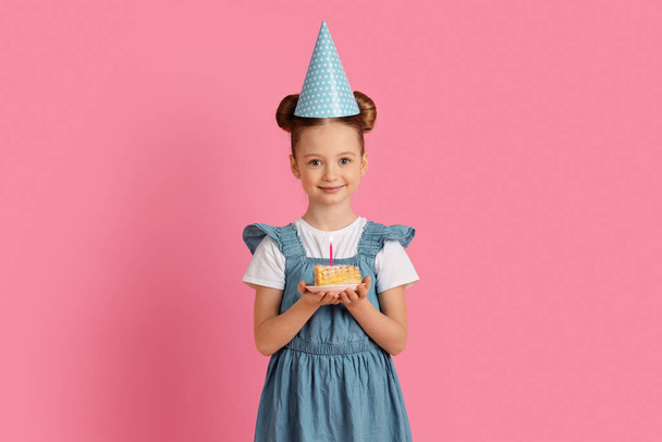 Ευτυχισμένο το μικρό κορίτσι στο κόμμα καπέλο κρατώντας κομμάτι της τούρτας γενεθλίων με κερί, χαριτωμένο χαμογελώντας Preteen θηλυκό παιδί γιορτάζει B-Day, Κάνοντας Ευχή, ενώ στέκεται πάνω από ροζ φόντο στούντιο, αντίγραφο χώρο - Φωτογραφία, εικόνα