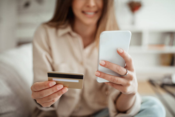Boldog Y-generációs európai női boltos hitelkártyával nézi a telefont a nappaliban, kivágva, bezárva. App for online shopping, finance banking, sales and cashback - Fotó, kép