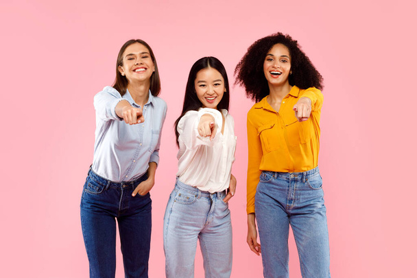 Hey, you. Portrait of happy multiethnic ladies in casual pointing fingers at camera and smiling, posing over pink background, studio shot - Photo, Image