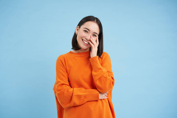 Portrait of young happy woman in orange jamper, touches her clear glowing skin, has beautiful white smile and candid emotions, blue background. - Foto, Imagem