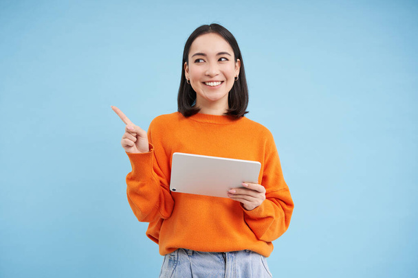 Modern asian woman with tablet, pointing at empty space for promo offer, banner, smiling and looking at advertisement, blue background. - Foto, Imagen
