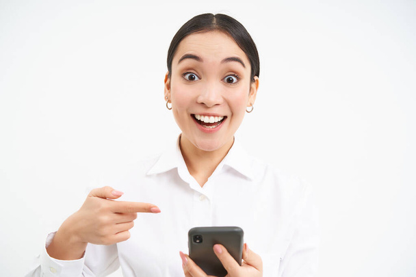 Крупный план портрет счастливой продавщицы, успешной женщины со смартфоном, указывая на свой телефон и улыбаясь удивлен, стоя на белом фоне. - Фото, изображение