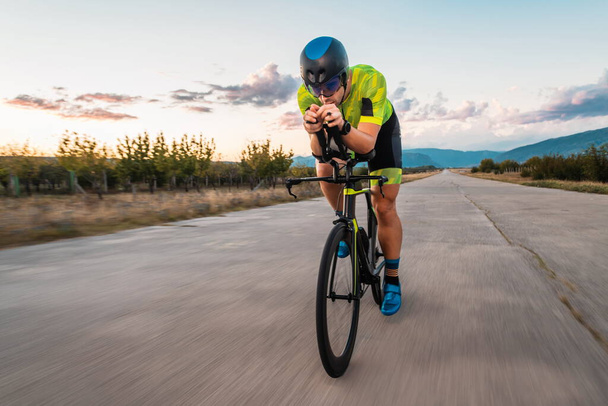  Triathlete riding his bicycle during sunset, preparing for a marathon. The warm colors of the sky provide a beautiful backdrop for his determined and focused effort - Foto, Imagen