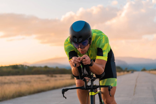  Triathlete riding his bicycle during sunset, preparing for a marathon. The warm colors of the sky provide a beautiful backdrop for his determined and focused effort - 写真・画像