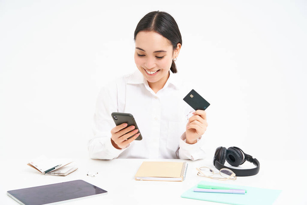 Şirket ve iş. Asyalı kadın ofiste, akıllı telefonu ve kredi kartı var, online ödeme yapıyor, doğrudan banka hesabı açıyor, beyaz arka plan. - Fotoğraf, Görsel