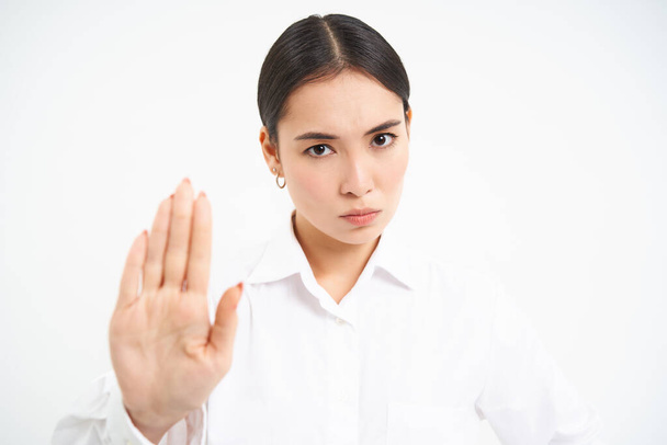 Будь ласка, зупиніться, досить. Серйозна азіатська жінка-менеджер, бізнес-леді показує розширений жест руки, не схвалює, забороняє smth, білий фон
. - Фото, зображення
