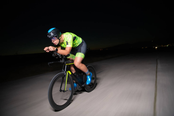 A triathlete rides his bike in the darkness of night, pushing himself to prepare for a marathon. The contrast between the darkness and the light of his bike creates a sense of drama and highlights the - Foto, Imagem