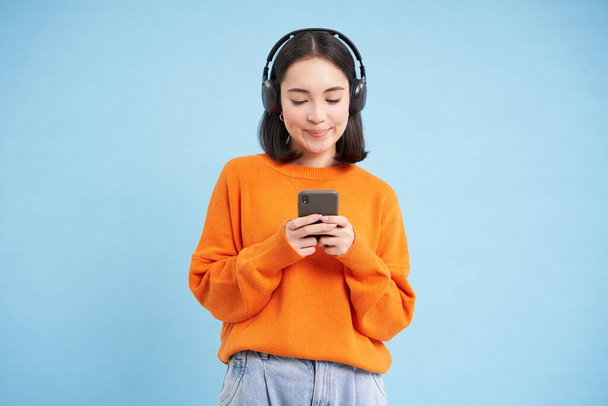 Szczęśliwa młoda, nowoczesna kobieta ze słuchawkami i smartfonem, słuchająca muzyki na telefonie komórkowym, cieszy się ulubionym podcastem, niebieskim tłem. - Zdjęcie, obraz