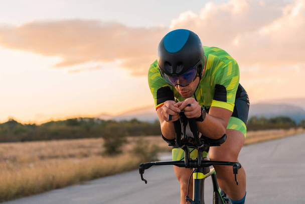 Triathlete riding his bicycle during sunset, preparing for a marathon. The warm colors of the sky provide a beautiful backdrop for his determined and focused effort - Foto, Imagem
