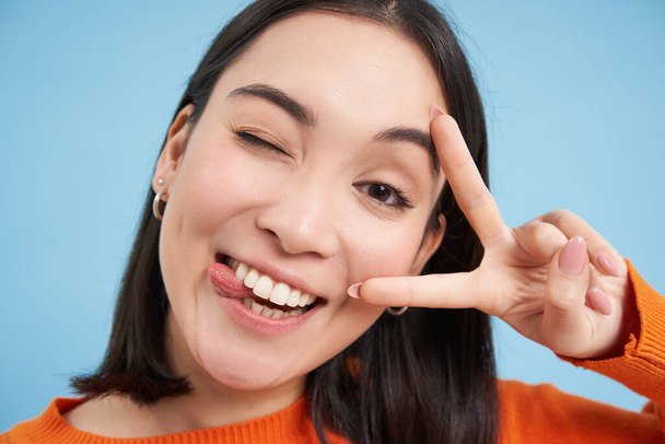 Close-up portret van positieve Aziatische vrouw, knipoogt en toont peave, v-teken gebaar, stokken tong en glimlacht, staat over blauwe achtergrond. - Foto, afbeelding