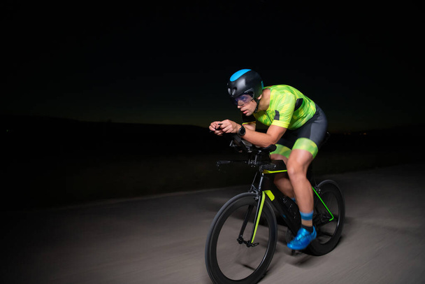 A triathlete rides his bike in the darkness of night, pushing himself to prepare for a marathon. The contrast between the darkness and the light of his bike creates a sense of drama and highlights the - Foto, imagen