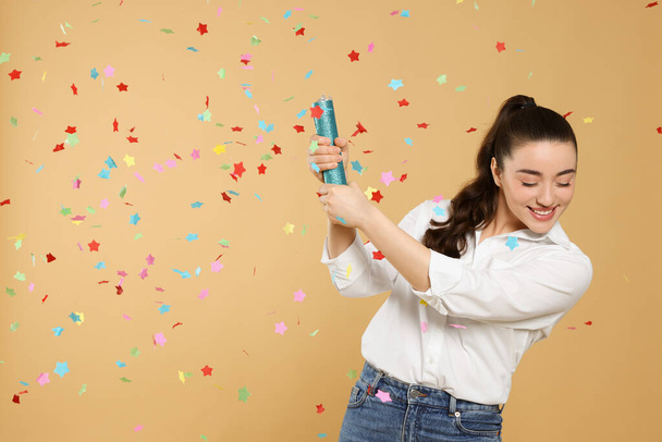 Young woman blowing up party popper on light brown background - Photo, Image