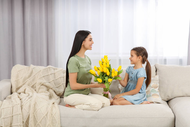 Happy woman and her cute daughter with bouquet of yellow tulips on sofa at home. Mother's day celebration - Foto, immagini