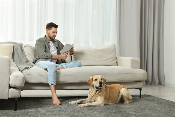 Man reading book on sofa near his cute Labrador Retriever at home - Photo, image