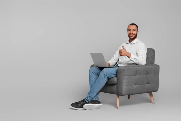 Smiling young man with laptop sitting in armchair on grey background. Space for text - Φωτογραφία, εικόνα