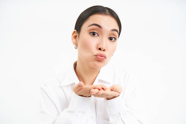 Close up portrait of cute office lady sends air kiss on palms, puckers lips and looks tender at camera, stands over white background. - Photo, Image