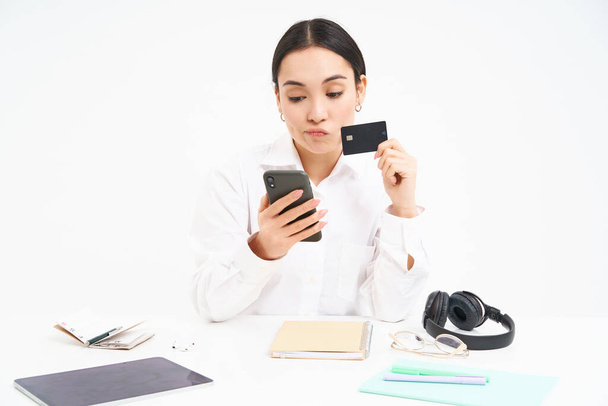 Junge Studentin sitzt mit Smartphone und Kreditkarte da, schaut schockiert auf ihr Bankkonto in der Handy-App, sitzt frustriert vor weißem Hintergrund. - Foto, Bild