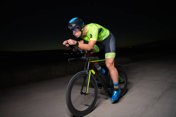 A triathlete rides his bike in the darkness of night, pushing himself to prepare for a marathon. The contrast between the darkness and the light of his bike creates a sense of drama and highlights the - Photo, Image