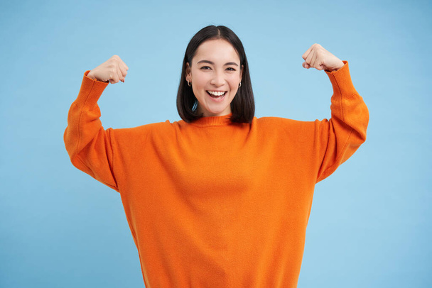 Des gens forts et en bonne santé. Jeune femme asiatique montre ses muscles, flexion biceps et semble confiant, souriant heureux, en grande forme, fond bleu. - Photo, image